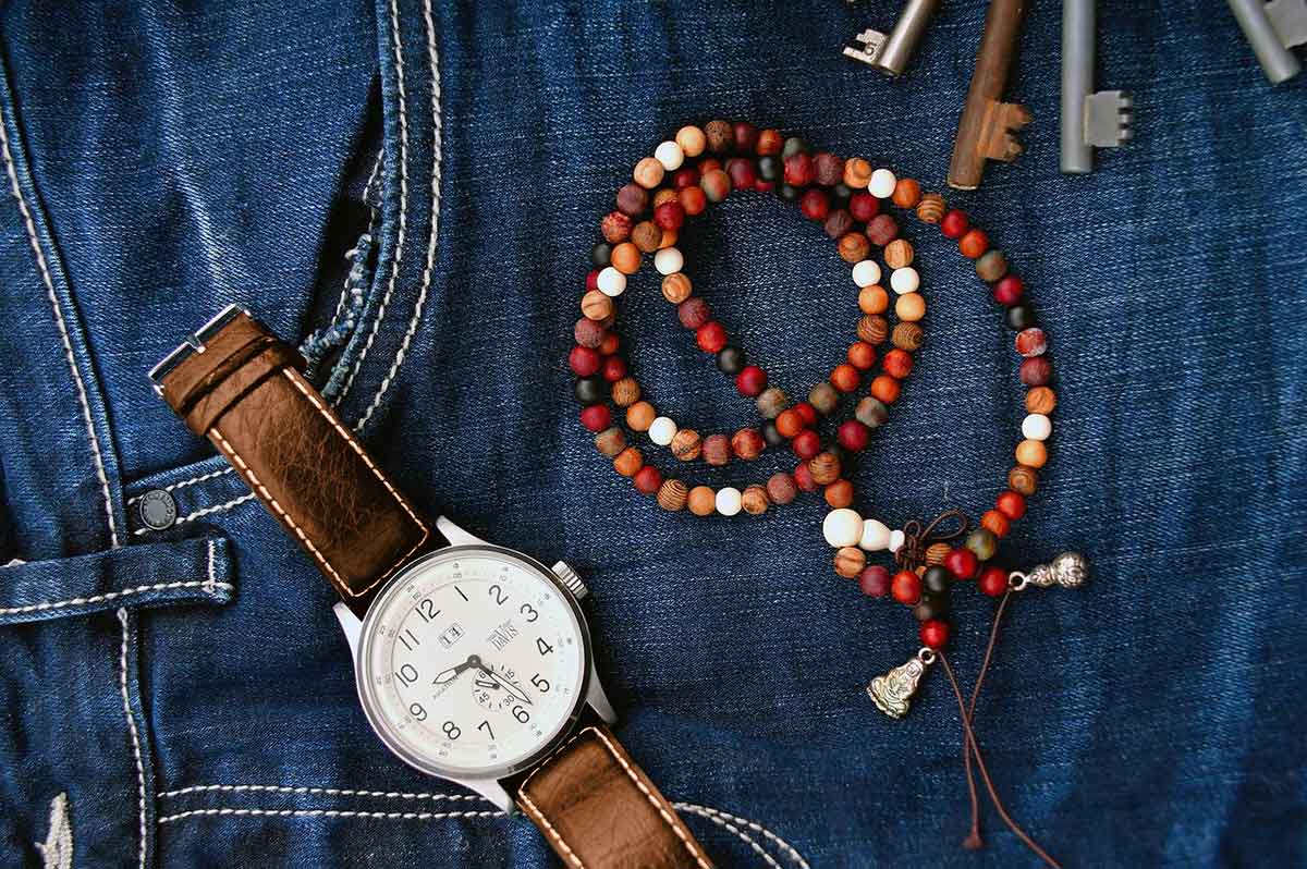 Brown leather watch and red, orange, and brown wood bead bracelet lying on blue jeans.
