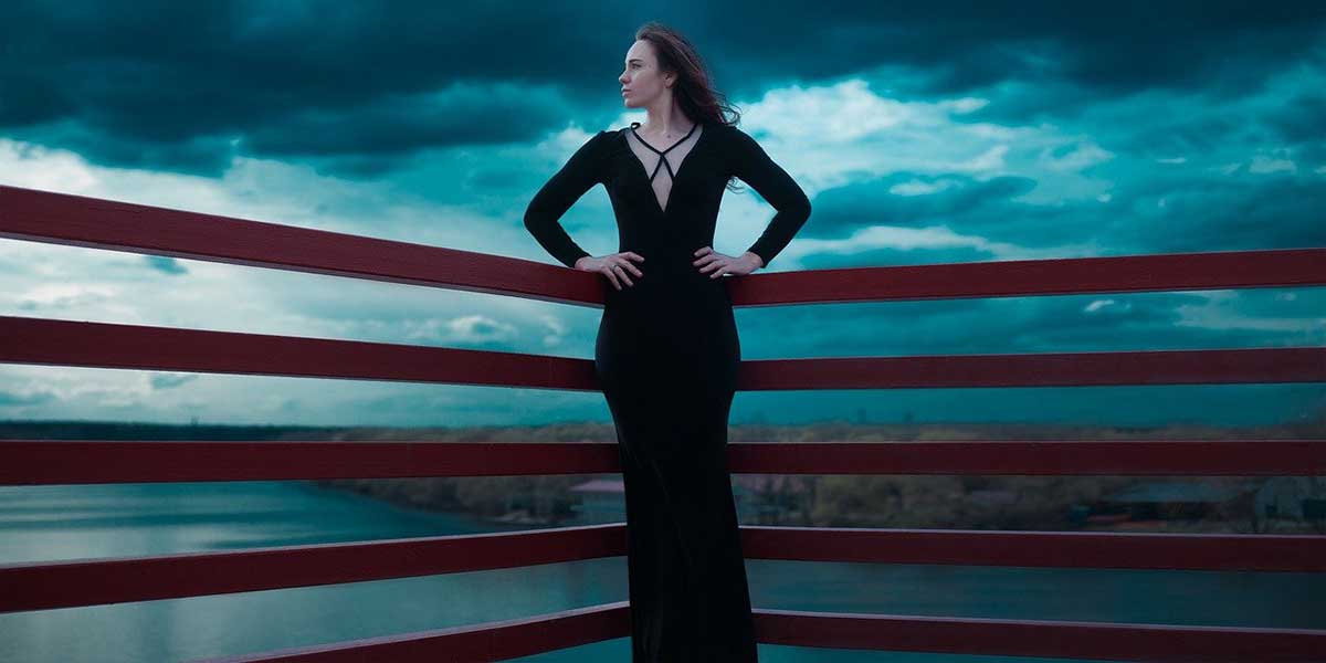 woman in elegant long black dress stands in front of dark moody skies.