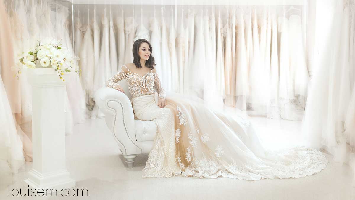bride dressed in white sits among white wedding dresses.